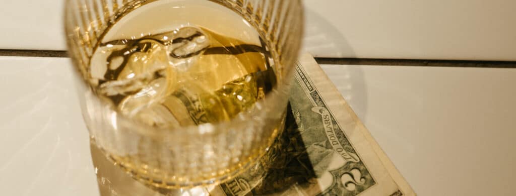 A glass of alcoholic drink on dollar bills, representing the connection between alcohol abuse and poverty
